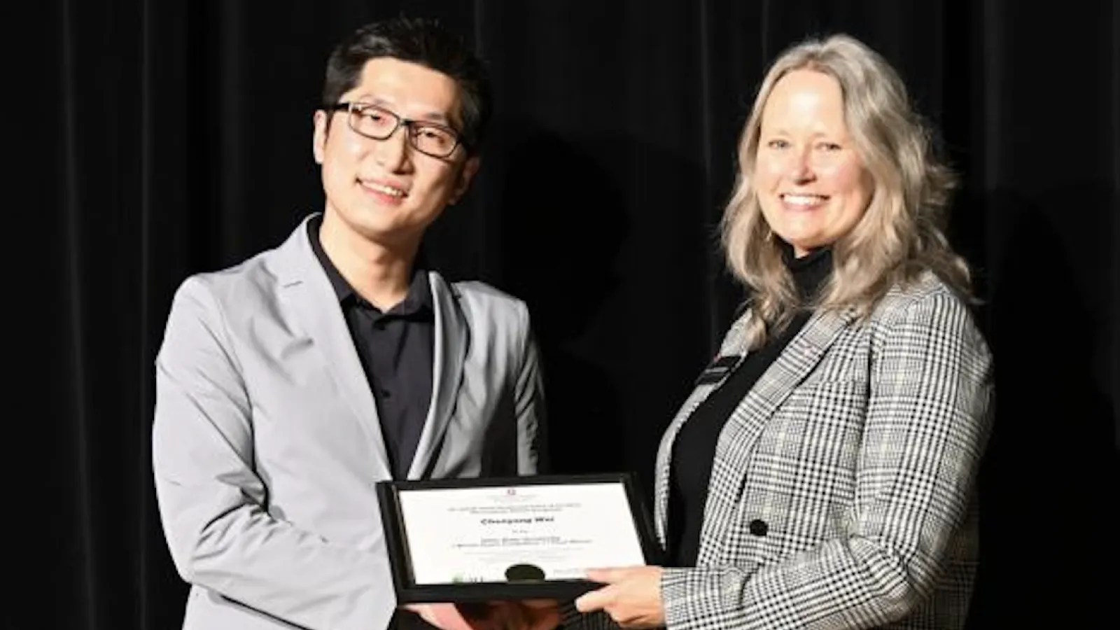Wei receiving certificate from Mary Stromberger in front of black background