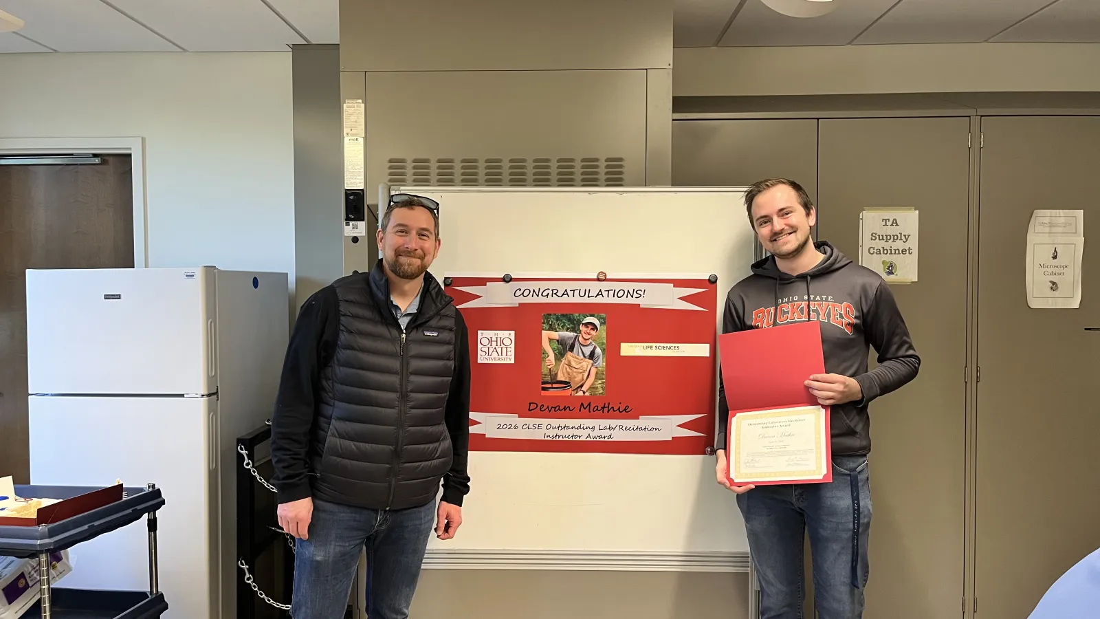 Jim Hood and Devan Mathie stand in front of poster that names Devan 2026 CLSE Outstanding Lab & Recitation Instructor
