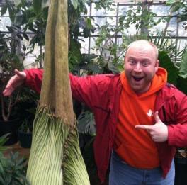 Macrander standing with right arm around spathe of Titan arum and left hand pointing to the plant