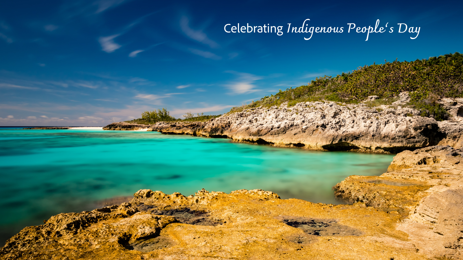 Half Moon Cay in San Salvador near Columbus' landing place. Celebrating Indigenous People's Day text in white over blue sky and water with rocky beach.