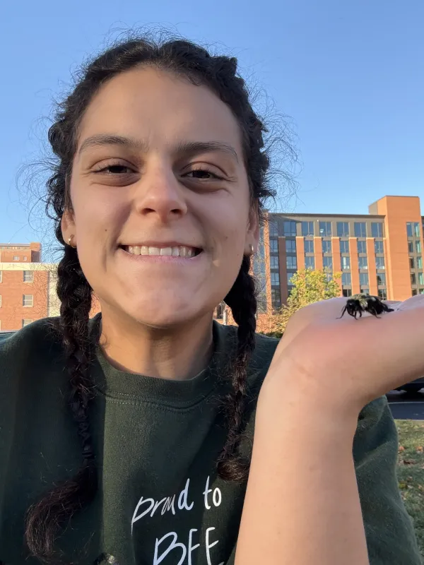 head and shoulders shot of Lizzy Sakulich holding bee in left hand in front of university buildings
