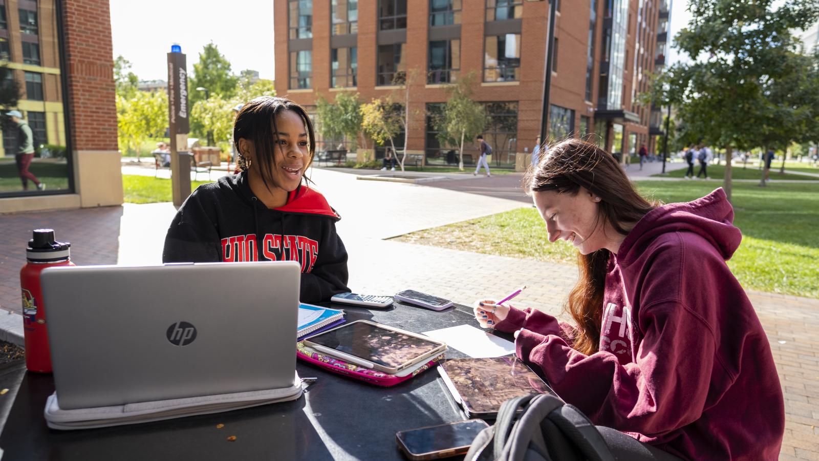 Two students working on electronic devices at table outside on campus